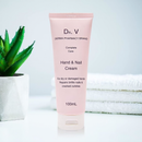 A tube of hand and nail cream from the Dr. V DERMA PHARMACY BRAND, placed on a white surface next to a potted plant and rolled up towels.