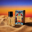 A perfume bottle and its packaging are placed on a sandy surface against a colorful sunset background.