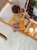 A wooden tray with a glass of red wine, a small box of vanilla and cardamom bath products, and two lit candles, all set on a white surface.