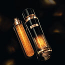 Two luxury perfume bottles with a golden glittery texture, one standing upright and the other lying on its side, against a dark background.