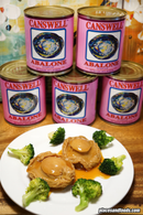 a plate of food with two pieces of meat covered in sauce, surrounded by broccoli and other vegetables. In the background, there are several cans of canned abalone, which appear to be the main focus of the image.