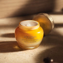 A jar of yellow cream with a white lid, labeled "L'OCCFTANE EN PROVENCE IMINOR TELLLE DIVINE CREME JEUNESSE" on a beige surface.