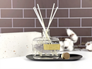 A clear glass bottle containing white reeds is placed on a black tray, with a white brick wall and some white cotton balls in the background.
