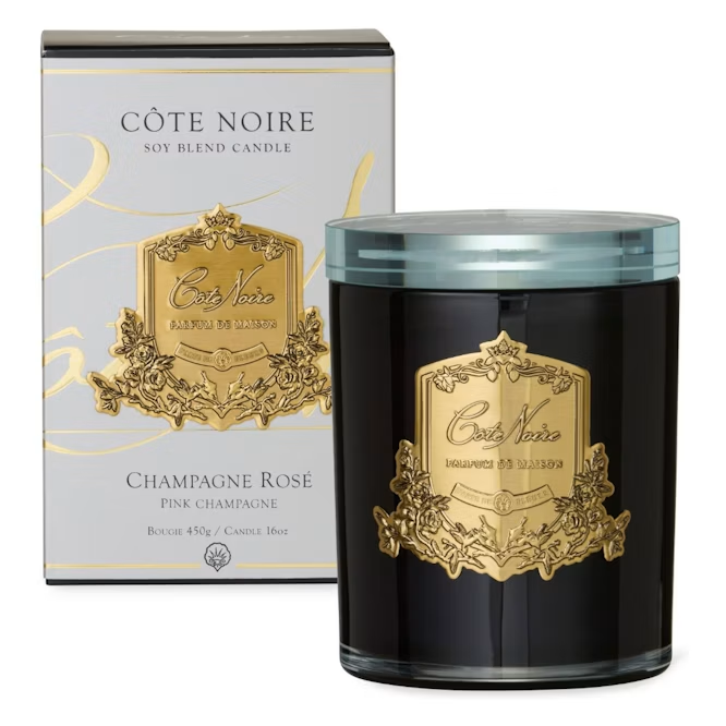 a Côte Noire soy blend candle and its packaging. The candle is a dark-colored, large candle with a gold label that reads "Champagne Rose" and "PINK CHAMPAGNE". The packaging is a light-colored box with a gold label that reads "Côte Noire SOY BLEND CANDLE".