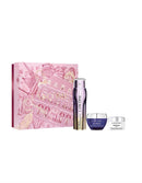 a collection of beauty products, including a pink box with a pink and gold design, a purple bottle, and two small jars, all arranged in a row.