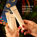 A person's hands holding two tubes of Jurique hand cream, with a background of a Christmas tree and a blue gift box.