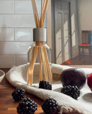 a glass jar filled with wooden sticks, a white cloth, and a few blackberries and an apple, all placed on a wooden surface.