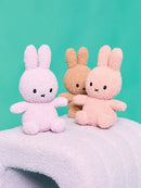 Three plush toy rabbits in different colors - pink, brown, and white - sitting on a white surface against a teal background.