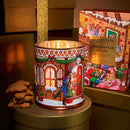 A decorative gingerbread house candle in a tin, with a stack of gingerbread cookies next to it, and a box of the same candle in the background.