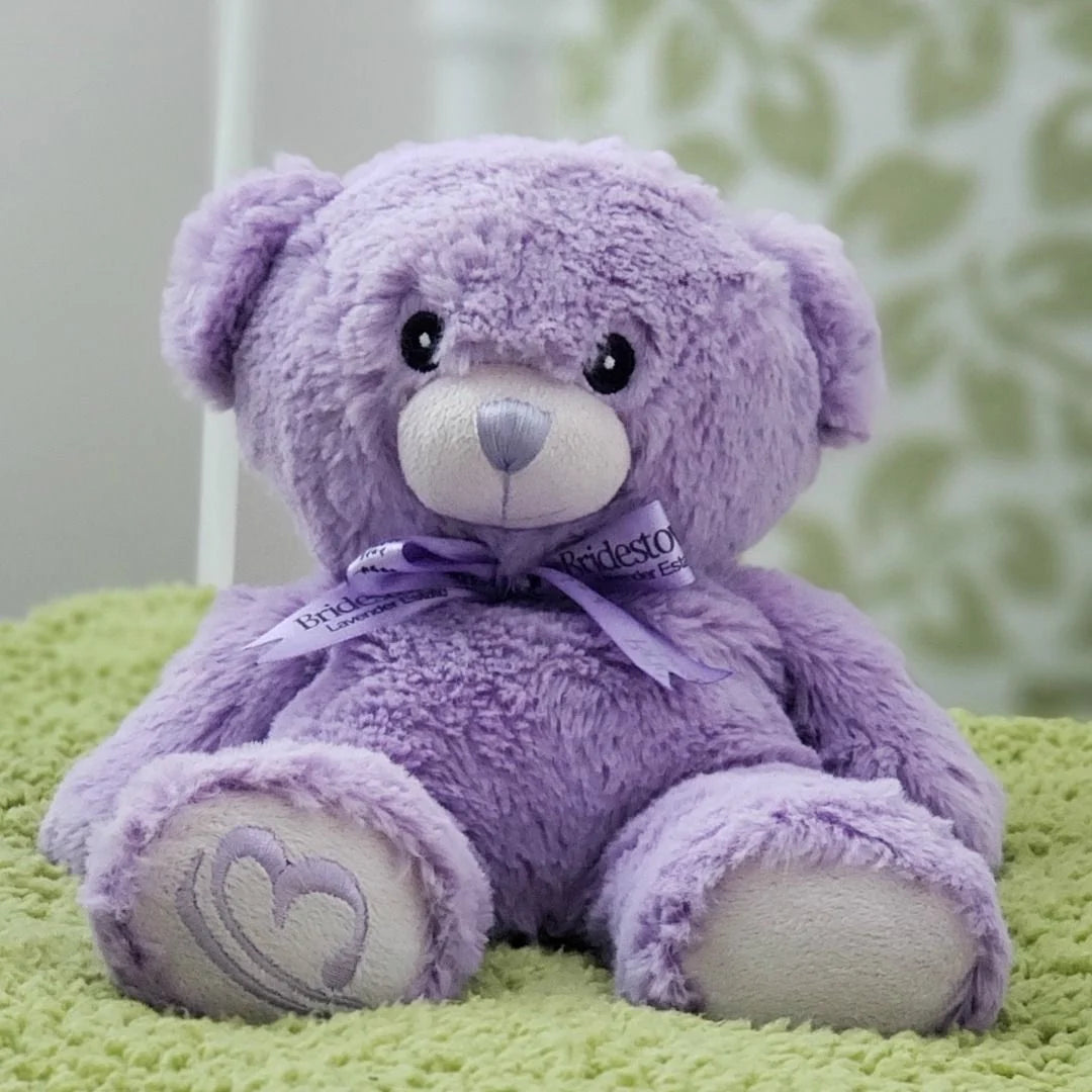 A purple teddy bear with a heart-shaped tag sits on a green surface, with a blurred green and white patterned background.