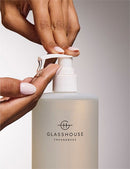 A person's hands are shown applying lotion from a transparent bottle with a pump dispenser, which is labeled "GLASSHOUSE Fragrances".