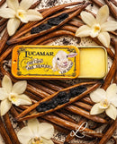a yellow tin of Lucamar lip balm surrounded by vanilla pods, blackberries, and white orchid flowers.