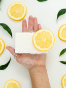 A hand holding a bar of soap with a slice of lemon on top, surrounded by lemon slices and green leaves.