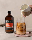 A hand is pouring a creamy beverage from a white ceramic pitcher into a glass with a swirling pattern, while a brown bottle labeled "MELROSE MCT" is placed next to the glass on a pink plate.