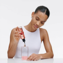 A woman in a white tank top is applying a pink liquid to a perfume bottle, with the bottle tilted towards her.