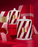 two large, striped candles with a red and white color scheme, placed on a red surface. The candles appear to be part of a holiday-themed collection, as suggested by the red and white striped boxes and the red background.