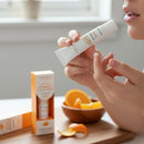 A person is holding a tube of vitamin C lip balm, with a wooden bowl of oranges and a bottle of the same product in the background.