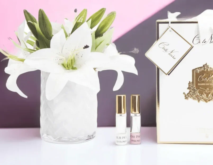 a bouquet of white lilies in a glass vase, along with two small perfume bottles, all placed on a white surface.