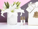 a bouquet of white lilies in a glass vase, along with two small perfume bottles, all placed on a white surface.