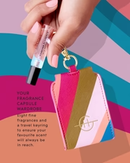 A hand holding a colorful keychain with a tube of lip balm attached to it, against a vibrant background.