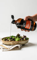 A hand is pouring a dark liquid, possibly a sauce or dressing, over a bowl of fresh salad greens.