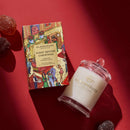 a box of Glashouse Fragrances Night Before Christmas Sugar Plums and a candle in a glass jar, both placed on a red surface.