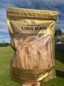 A hand holding a bag of dried fish, specifically Chinese dried fish, against a backdrop of a blue sky and green grass.