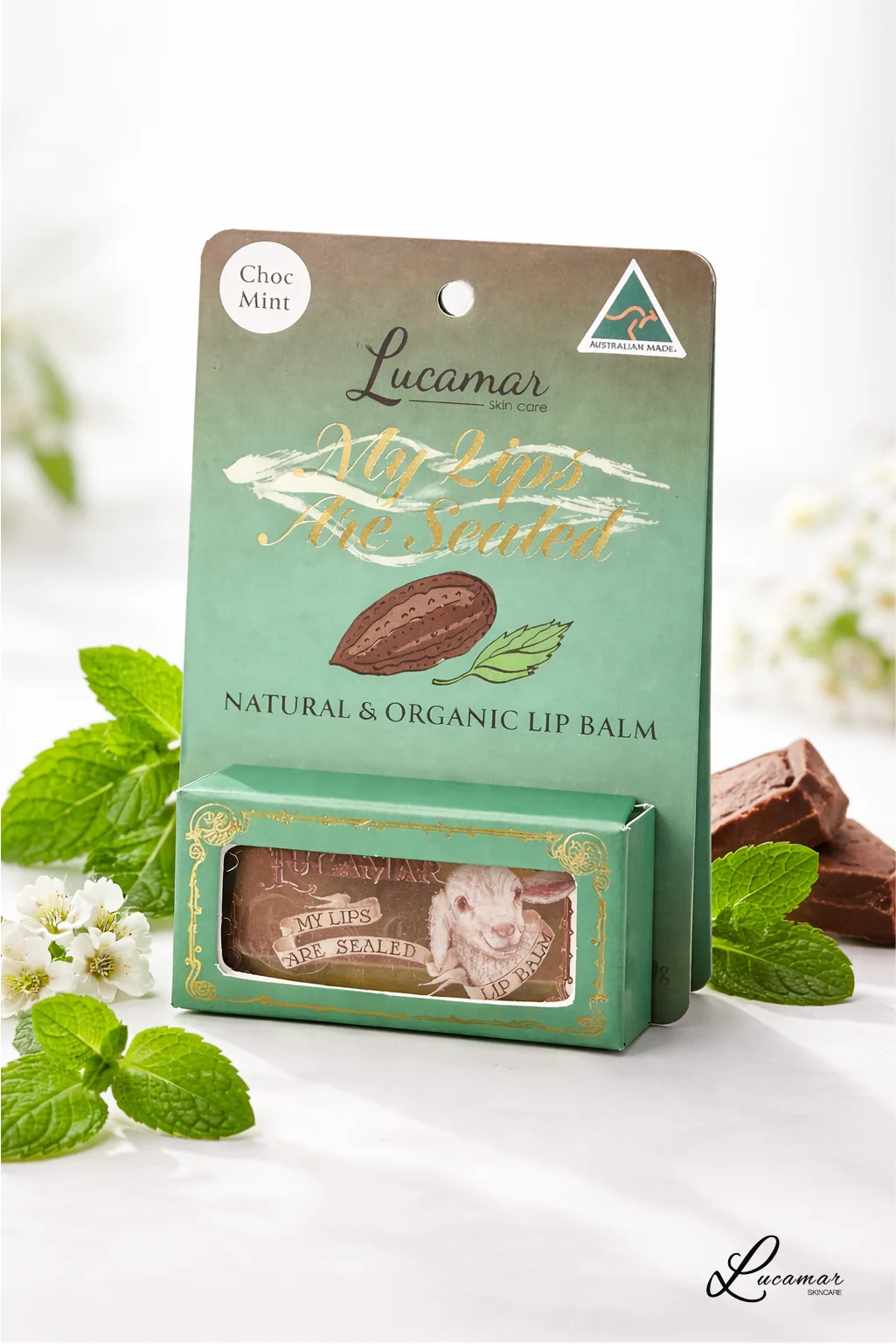 a product called "My lips are sealed" by Lucamar, a natural and organic lip balm. The product is displayed in a green box with a picture of a chocolate bar and mint leaves. The background includes a blurred white background with some flowers.
