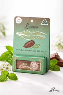 a product called "My lips are sealed" by Lucamar, a natural and organic lip balm. The product is displayed in a green box with a picture of a chocolate bar and mint leaves. The background includes a blurred white background with some flowers.