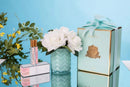 a collection of beauty and personal care products, including a vase with white roses, a box, and a bottle, all set against a blue background.