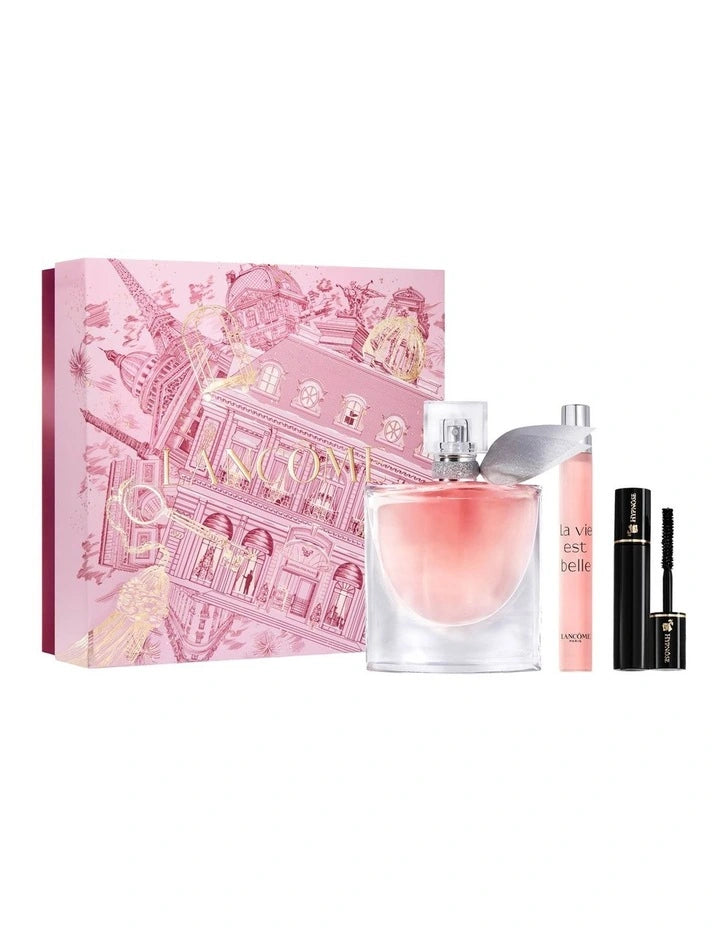 a pink box with a pink and white illustration of a building, along with a clear perfume bottle and a black mascara tube.