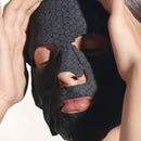 A close-up of a person's face wearing a black hexagonal patterned mask, with their hands covering their face.