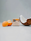 a box of coconut and finger lime balm, along with a partially peeled coconut and an orange peel, all placed on a white surface.