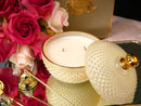 a white candle in a glass bowl, surrounded by a bouquet of pink roses and a gold-colored box.
