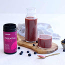 a bottle of melrose essential red supplement, a glass of red smoothie, and a wooden cutting board with blueberries, a spoon, and a bowl of blueberries.